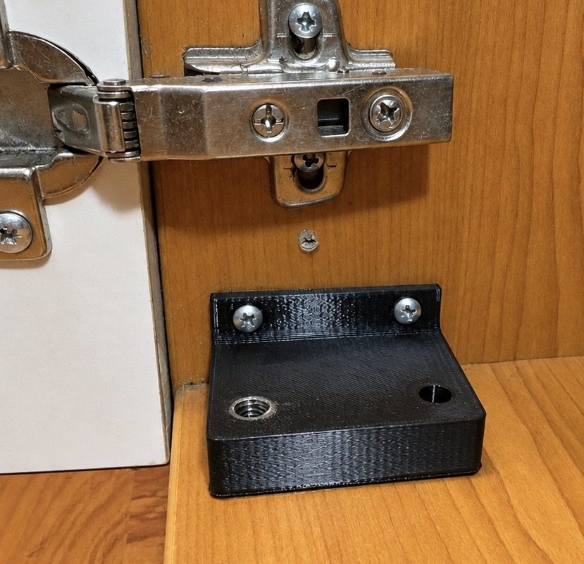 Černý 3D tištěný díl namontovaný pod pantem skříňky